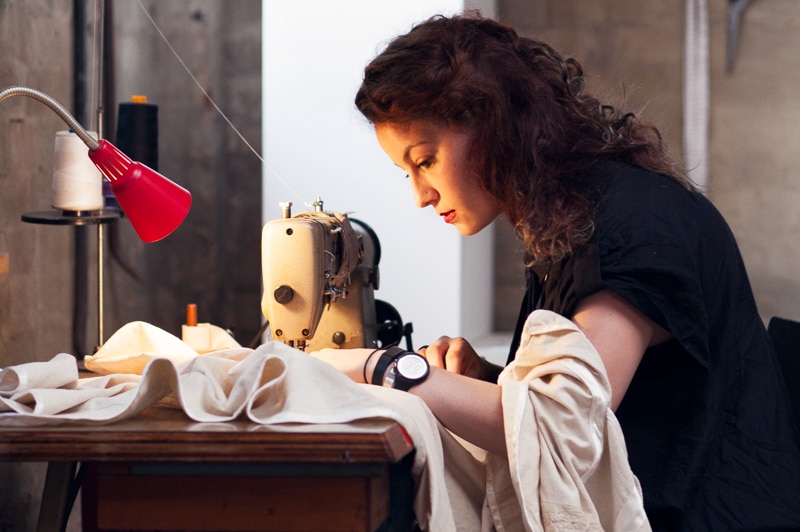 Fashion designer Pia Interlandi sits at table using sewing machine to make funeral shroud.