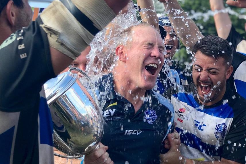 A group of men wearing striped jerseys surround a man holding a large trophy as water is poured over them all.