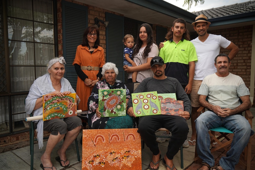 An elderly woman sits with her family who are holding up traditional Indigenous dot paintings.