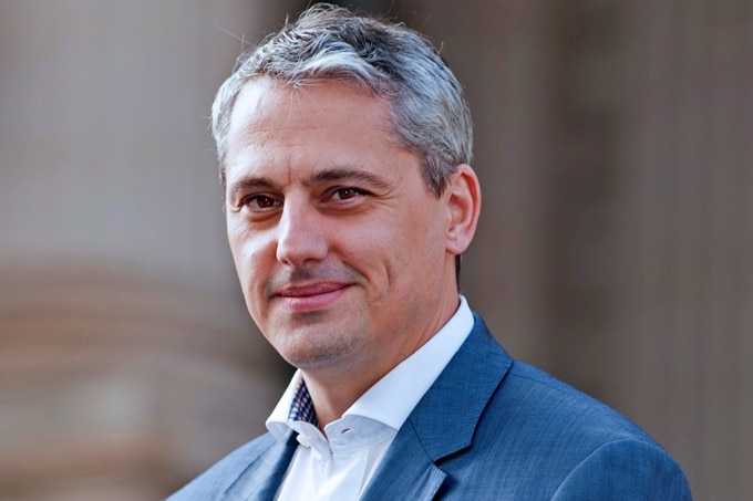 A portrait of a man with silver hair wearing a suit jacket and no tie.