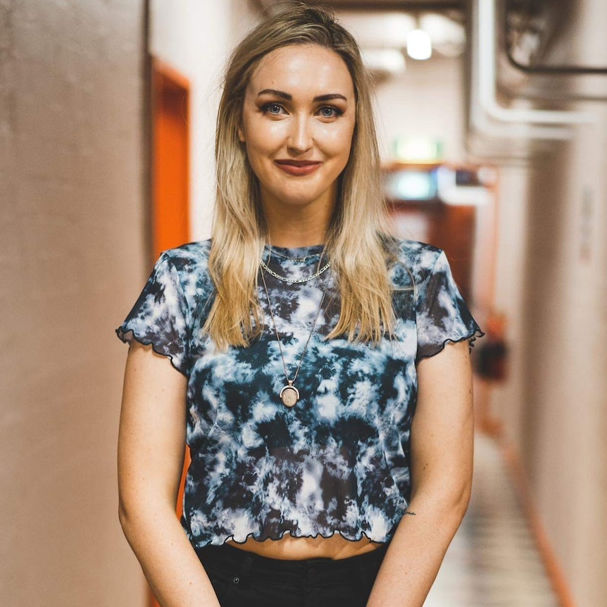 Joey wears a blue tie-dyed shirt and makeup, standing in a hallway smiling.