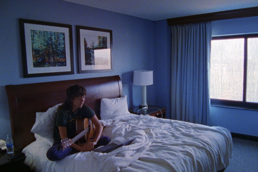 A white woman with shaggy brown hair wears a dark t-shirt and sweatpants and plays guitar on a bed in a blue bedroom.