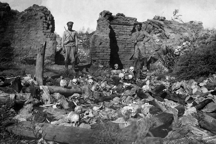 A black and white photo shows soldiers standing in ruins before a sea of skulls and bones in front of them.