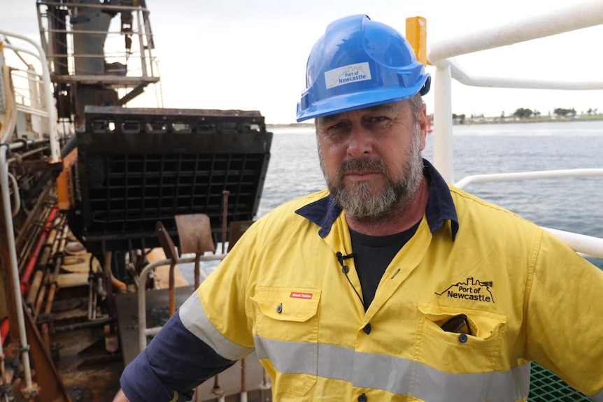 A man with a beard and a blue hard hat and high vis clothes with a dredge pipe in the background.