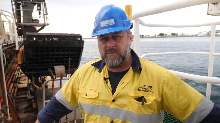 A man with a beard and a blue hard hat and high vis clothes with a dredge pipe in the background.