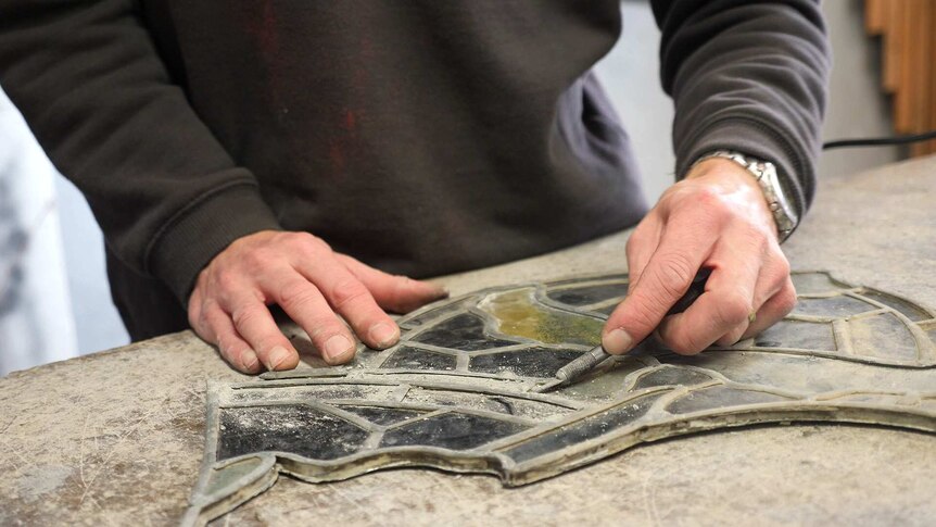 A glass restorer uses a scalpel to carefully scrape dirt from an ancient piece of window
