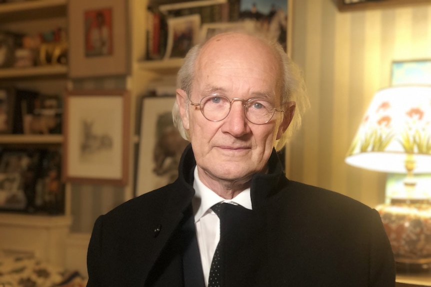 An older man wearing spectacles and a dark coat in a living room.