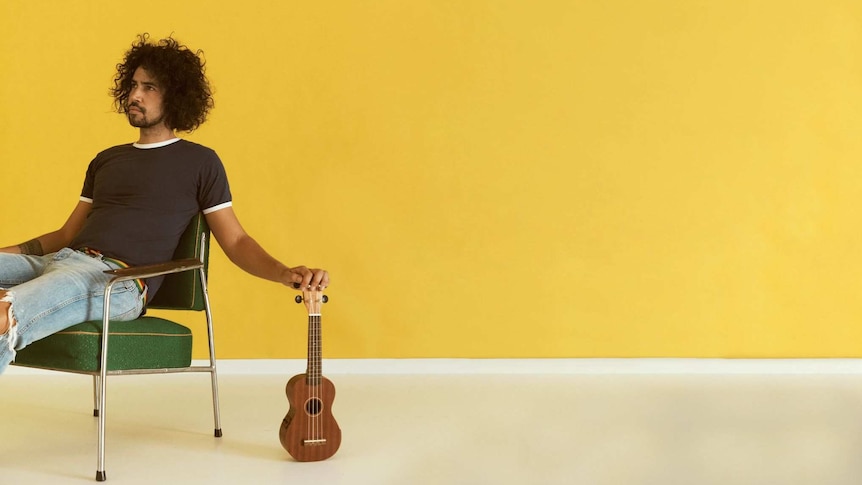 Press shot for Bobby Alu. He sits in a vintage chair to the left of a ukulele which his hand is resting on.