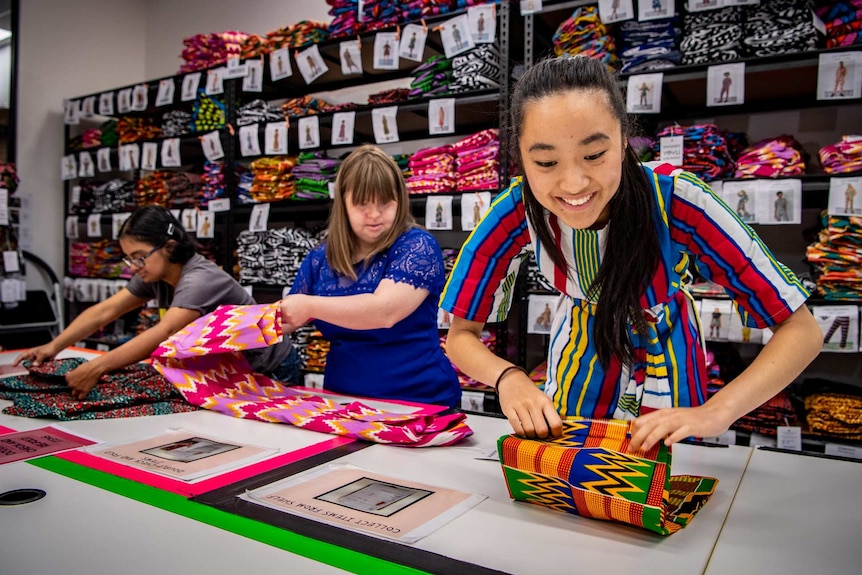 Three women fold clothes in  front of shelves of folded up brightly coloured clothing.