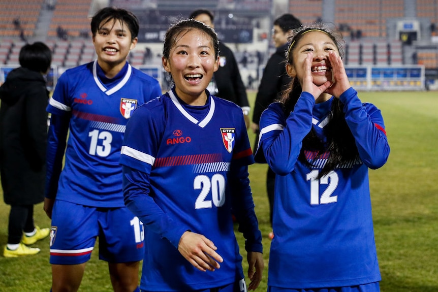 Three footballers smile towards a crowd in a stadium