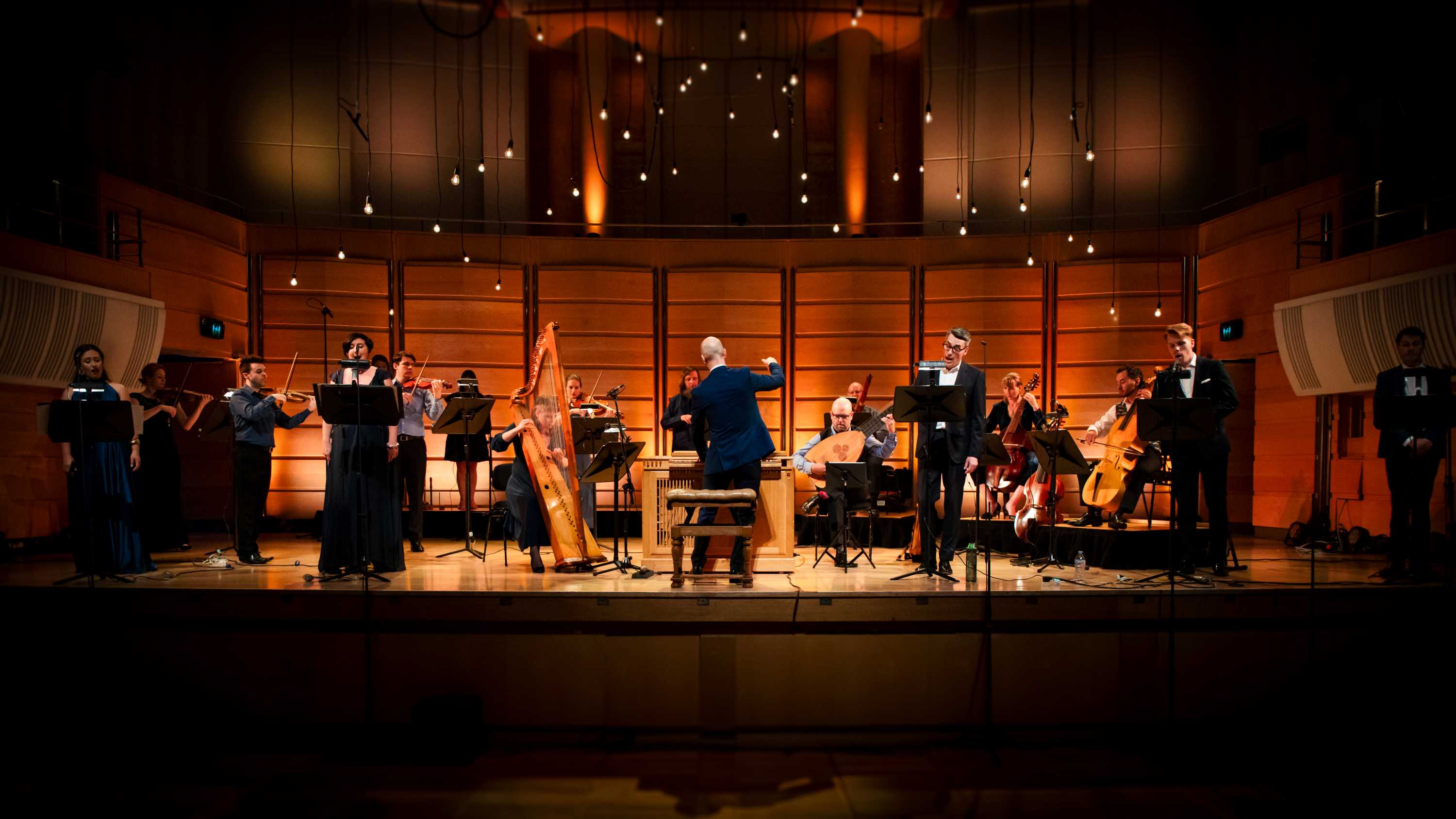 An early music ensemble on stage in smart casual clothes, with five vocal soloists in formal wear standing in front.