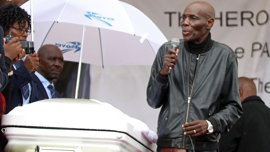 A man in a leather jacker and jeans holds a microphone to his mouth and purses his lips as he sings at a funeral.