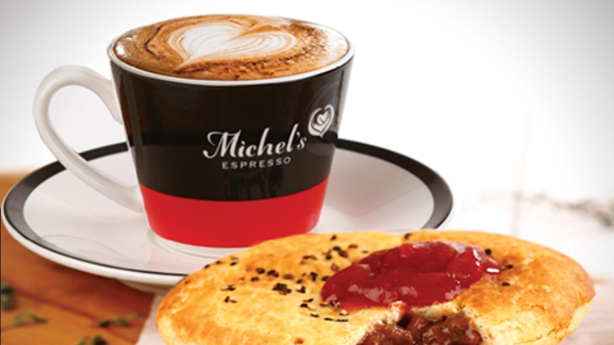 A coffee in a Michel's Espresso mug and a meat pie with tomato sauce on top sitting on table