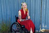 A blonde woman in a bright red dress, in a wheelchair, smiles widely