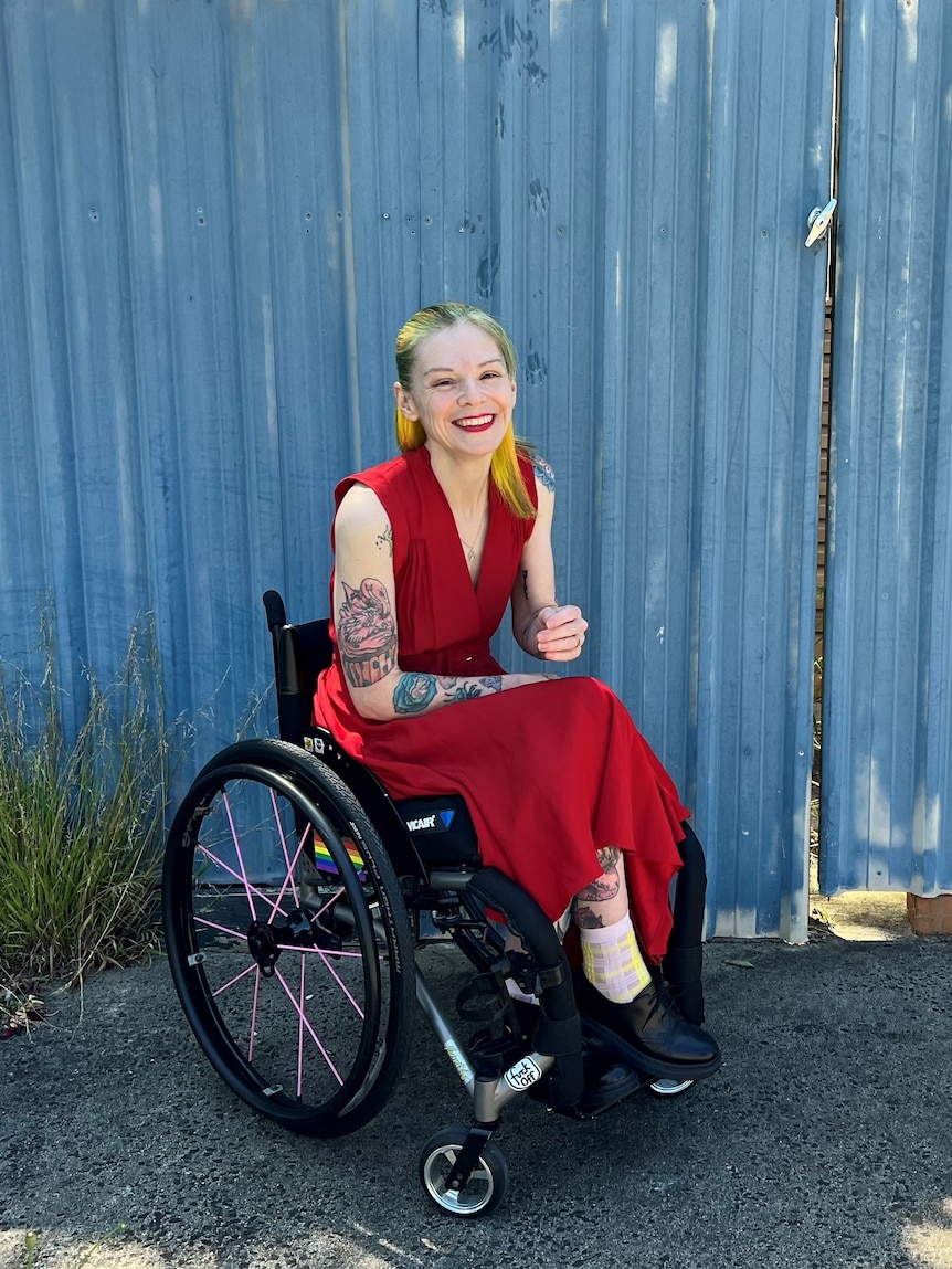 A blonde woman in a bright red dress, in a wheelchair, smiles widely
