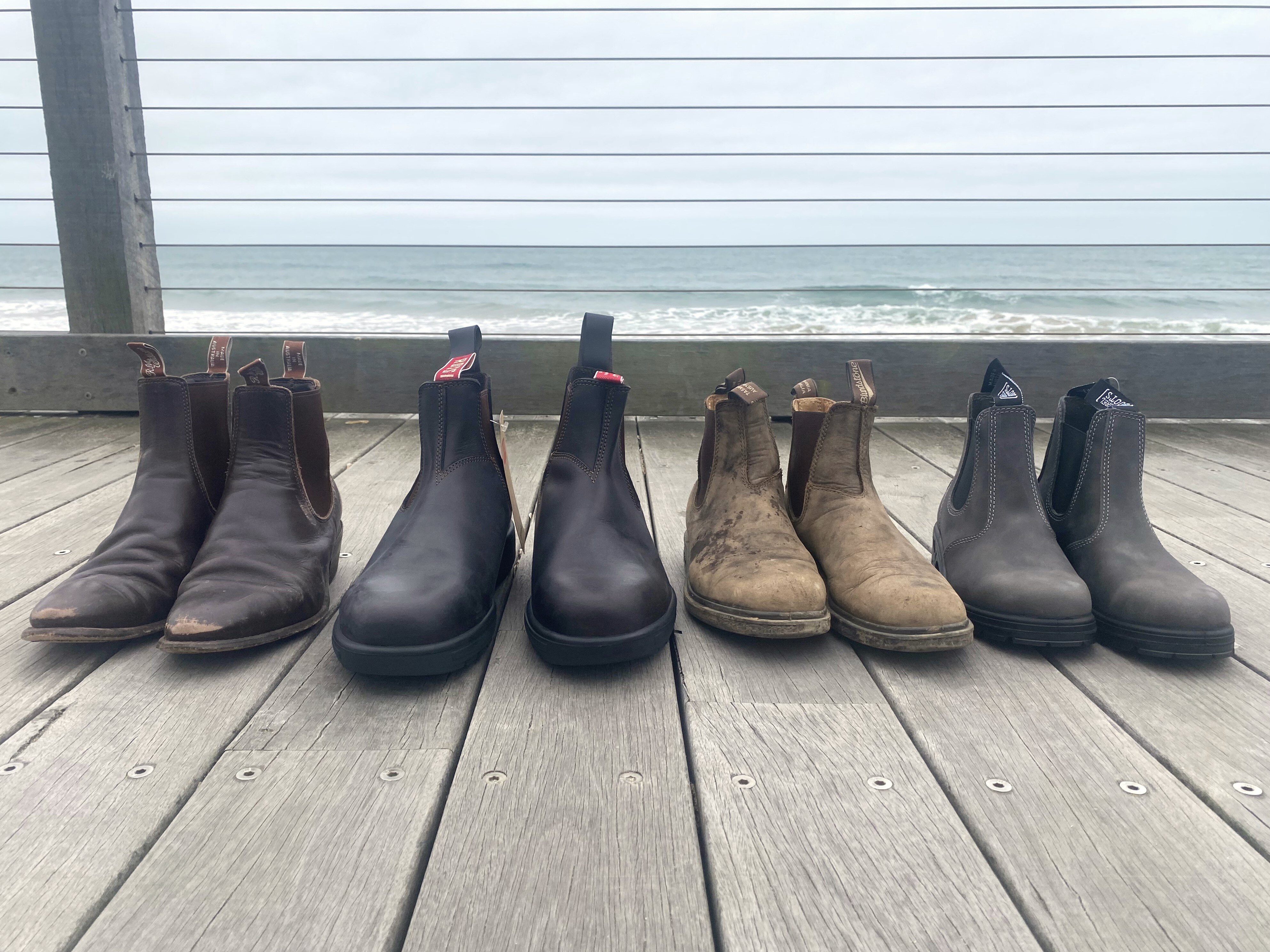 Four pairs of leather boots lined up on wooden boardwalk with ocean in background