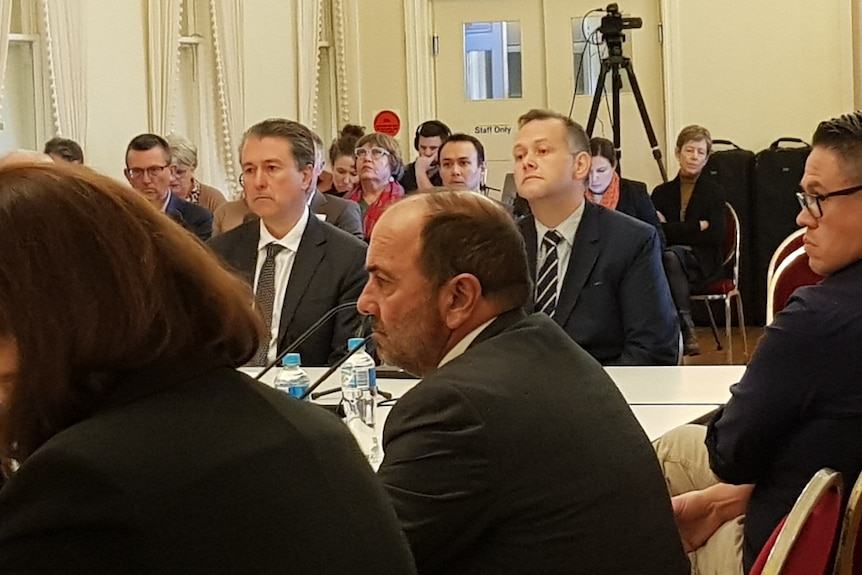 Two men sitting at a desk, with people in a public gallery behind them. 