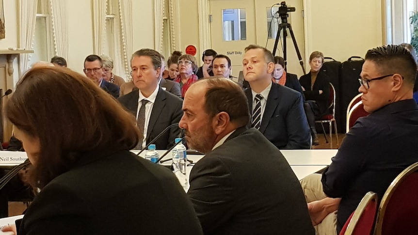 Two men sitting at a desk in an inquiry with people in a public gallery behind them. 