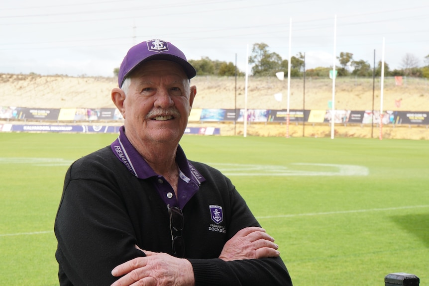 A man wearing a Fremantle Dockers hat and shirt stands with his arms folded