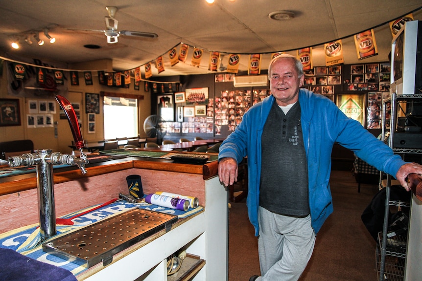 A man standing in a country pub's bar, with tables and a TV behind him, and walls covered in photos.