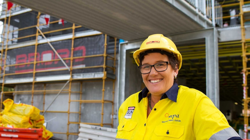 A woman, wearing a yellow hard hat and a yellow high-vis shirt, stands in front of a construction site.