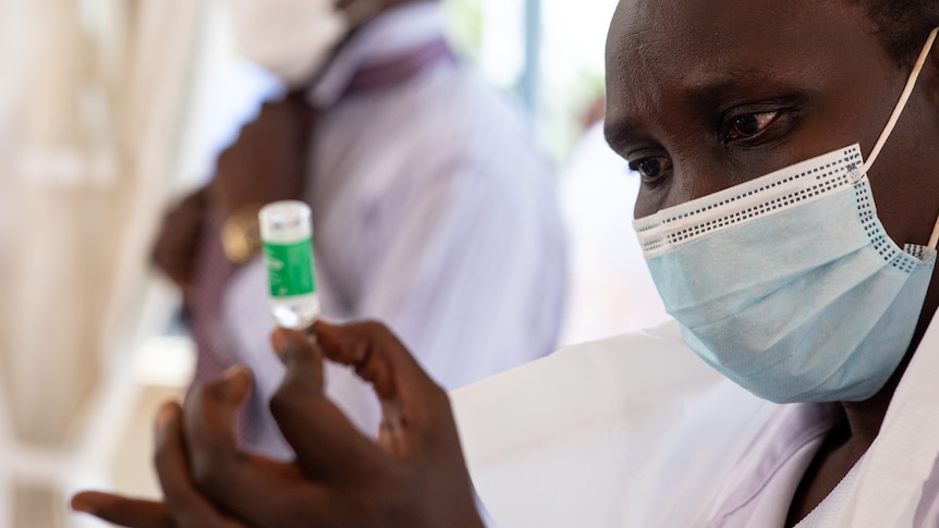 An African woman in a surgical mask holding a small vial between finger and thumb