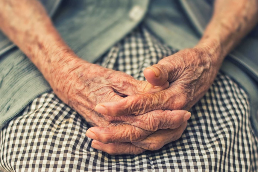 An elderly woman sits with her hands in her lap.