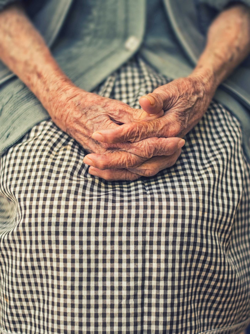 An elderly woman sits with her hands in her lap.