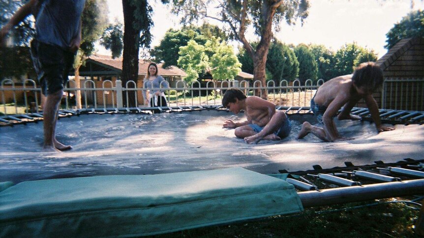 Three young children jump on trampoline with young girl looking on in background