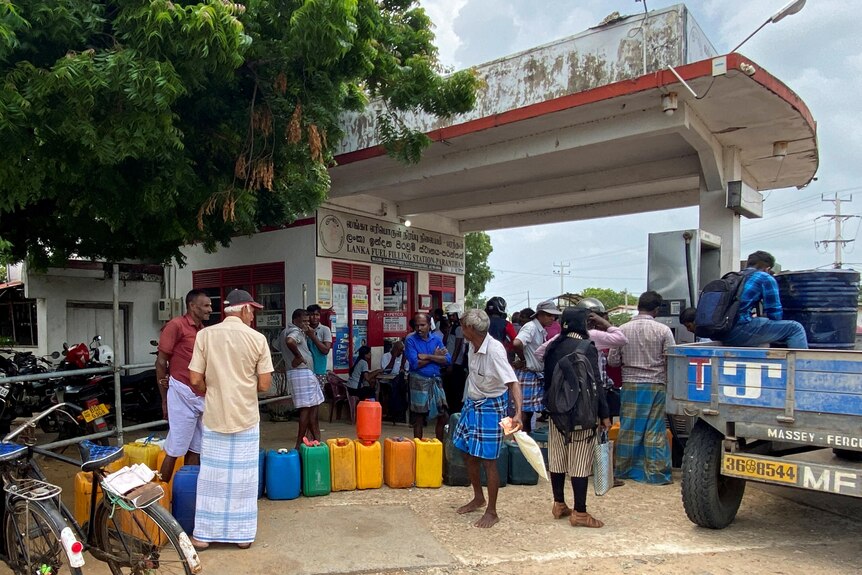 Hombres de Sri Lanka hacen fila frente a una gasolinera, con contenedores de combustible multicolores esperando.