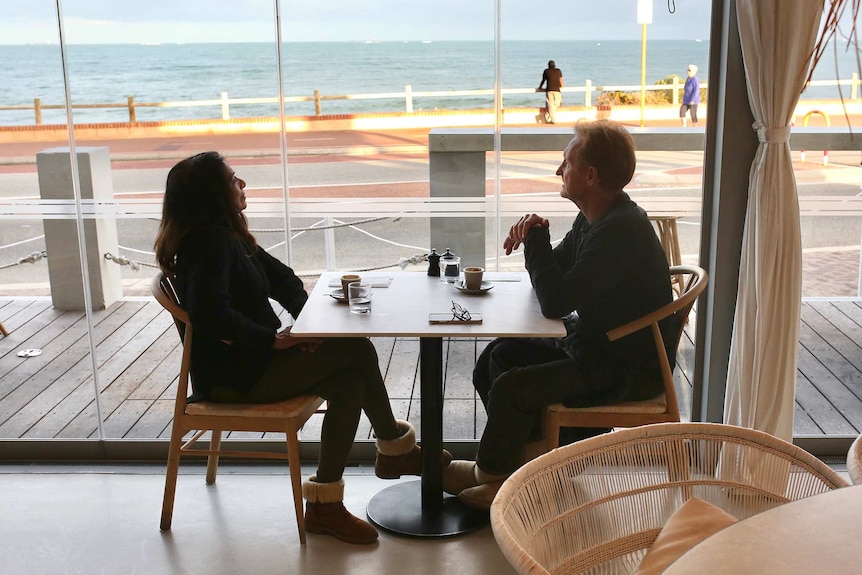 A man and woman sit at a table in a cafe next to windows looking out over the beach in Perth.