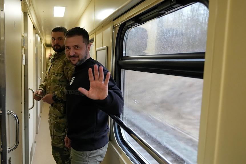 A man in a train corridor waves to the camera.
