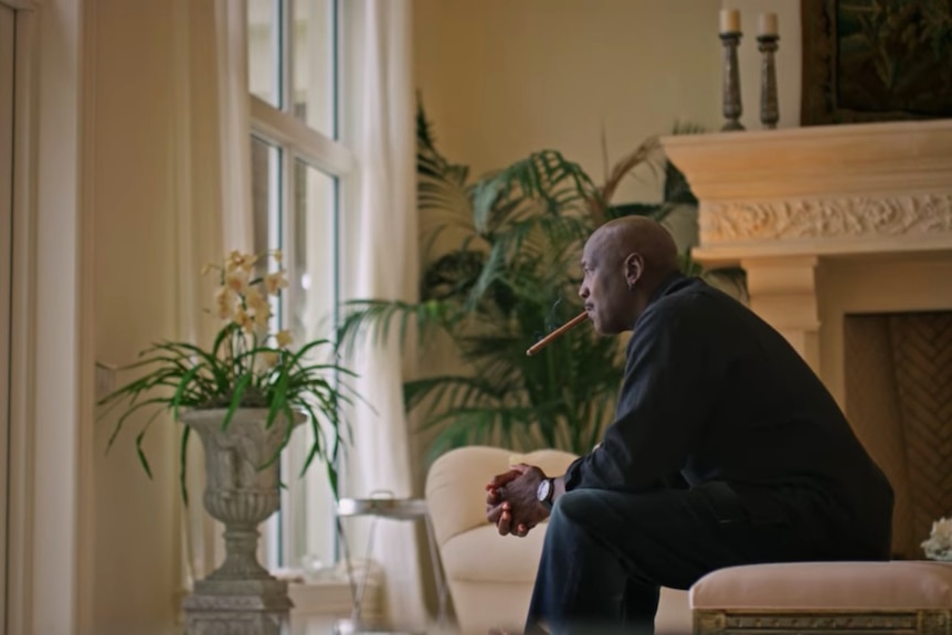 A seated Michael Jordan looks out a window while smoking a cigar during the documentary The Last Dance.