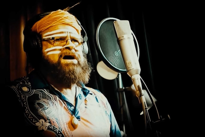 An Aboriginal man with face paint and a beard sings into a microphone