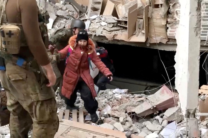 A child in pink jacket climbs over rubble towards a soldier. 