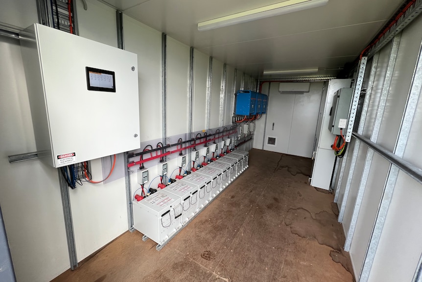 The inside of a shipping container with batteries wired together on the floor, surrounded by electrical equipment.