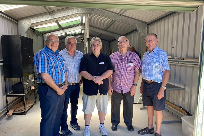 Five older men stand in a shed, smiling.