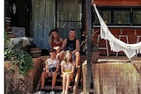 Man and woman sit on steps in front of a house with two children sitting in front of them. A white hammock hangs by their side. 