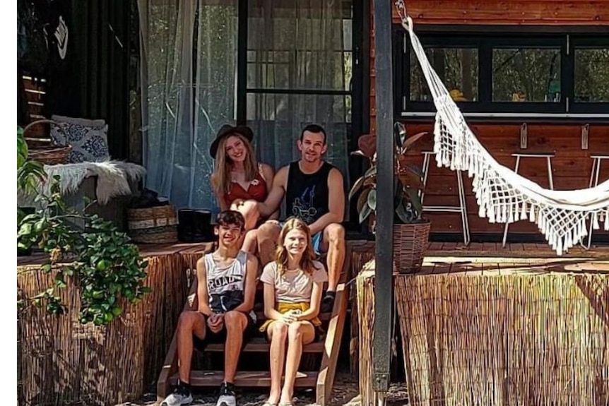 Man and woman sit on steps in front of a house with two children sitting in front of them. A white hammock hangs by their side. 