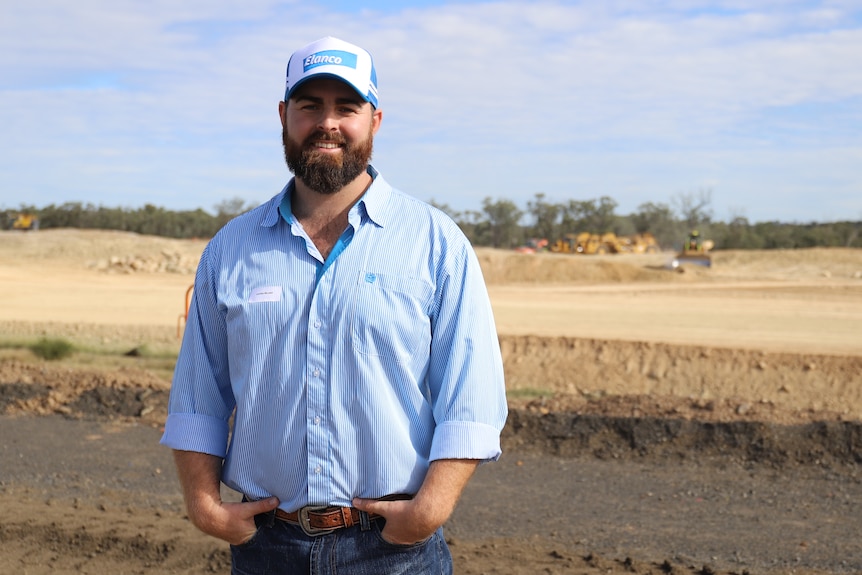 Man with blue shirt and a cap on stands in front of cleared land with his hands in his pockets. 