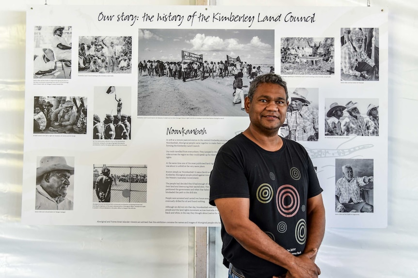 Anthony Watson stands in front of a board with pictures and words detailing the history of the Kimberley Land Council.