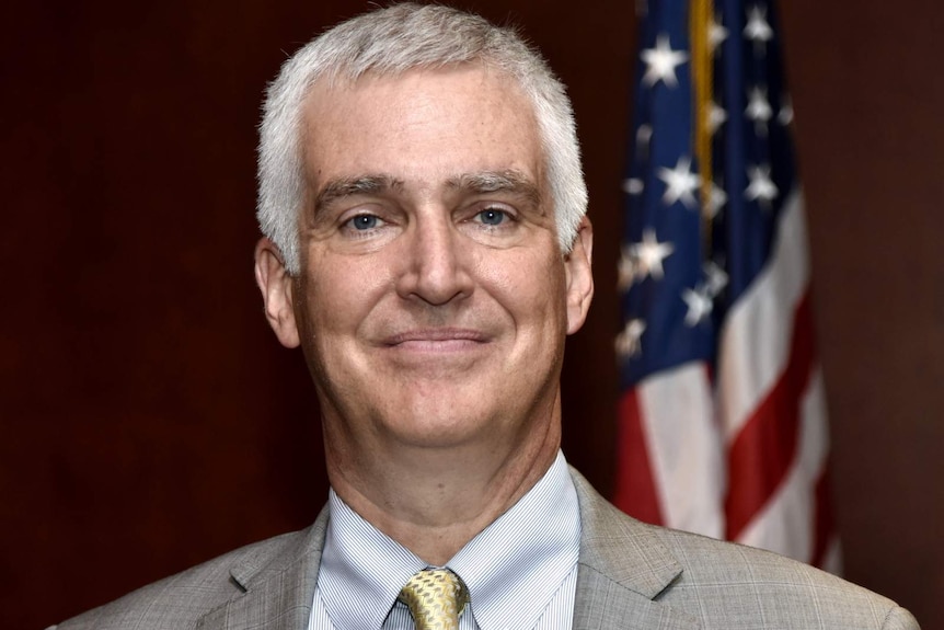 Fred Fleitz smiling, he is wearing a grey suit and is standing in front of an American flag.