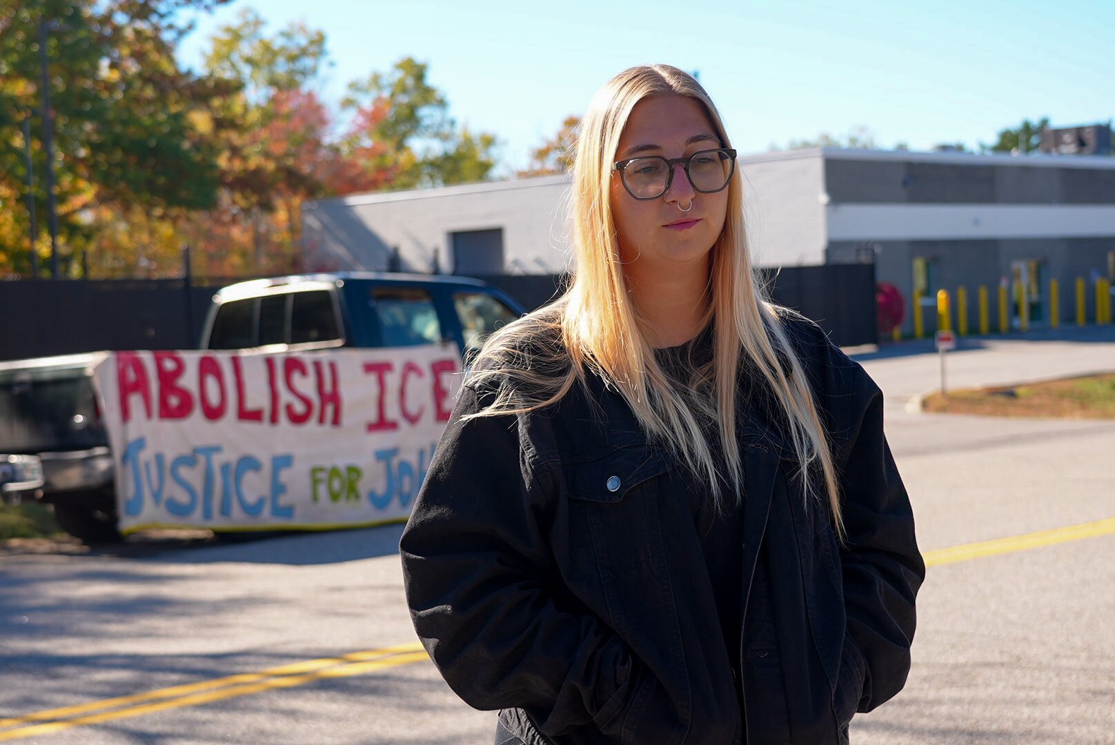A blonde woman in a black jacket. Behind her is a banner saying 'Abolish ICE' and a building.