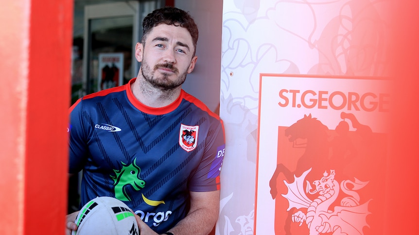 Adam Clune stands next to a St George Illawarra sign holding a football.