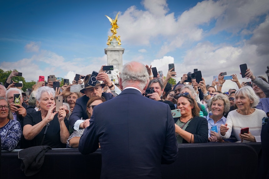 King Charles with his back to the camera, facing a large crowd of people holding phones 