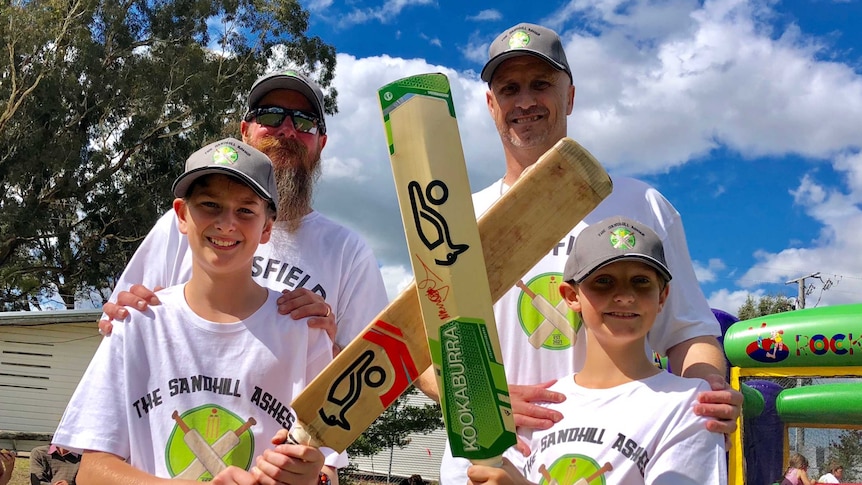 Two men stand behind their two young sons holding cricket bats.