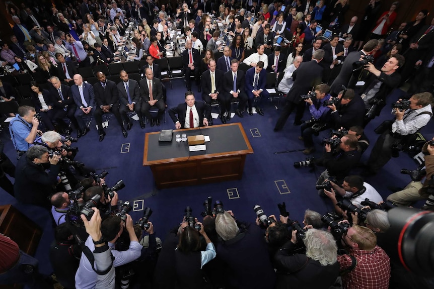 James Comey takes his seat surrounded by photographers and others.