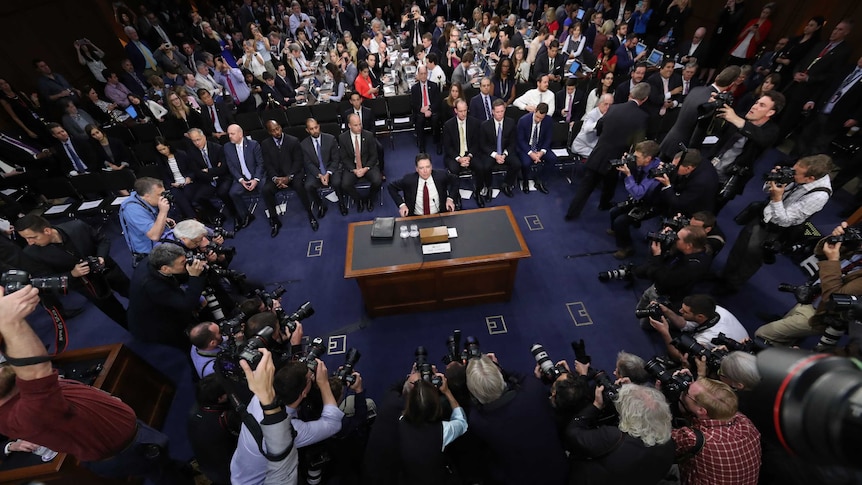 James Comey takes his seat surrounded by photographers and others.