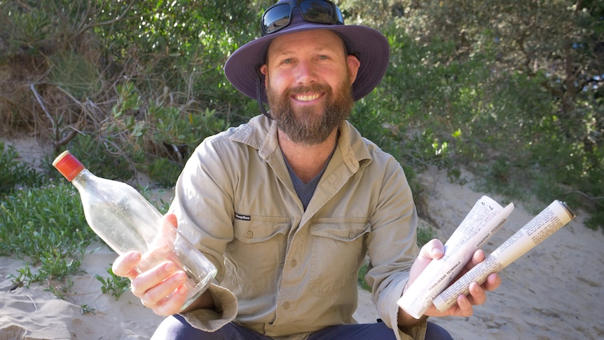 Portrait of man in outdoor work gear crouching on beach holding an empty bottle and rolled up message