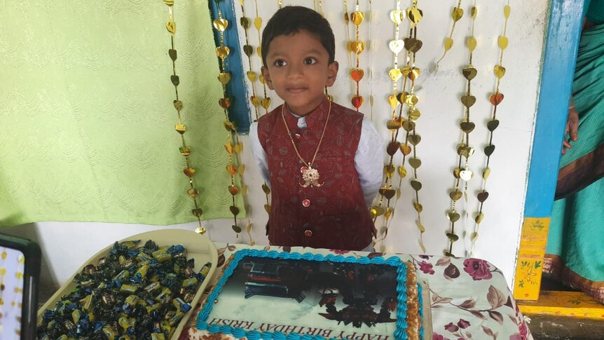A little boy in a vest standing behind a cake reading "happy birthday Krish"